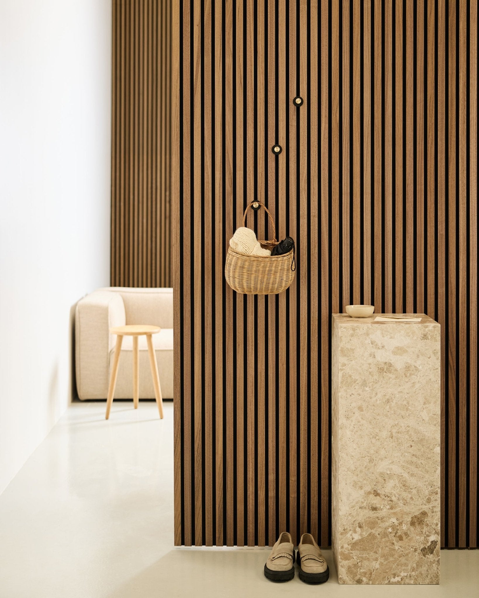 Entrée minimaliste avec mur à lattes acoustiques Akupanel, patère WoodUpp Norr en black et brass, panier, piédestal en pierre, chaussons et chaise beige.